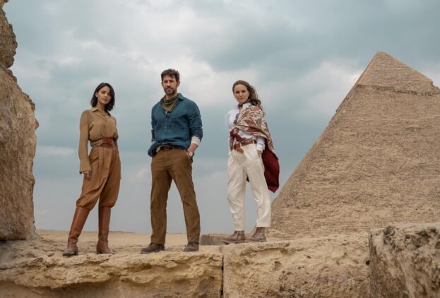 Three people stand on stone ruins near a pyramid in Egypt, dressed in casual, earth-toned clothing, under a cloudy sky.